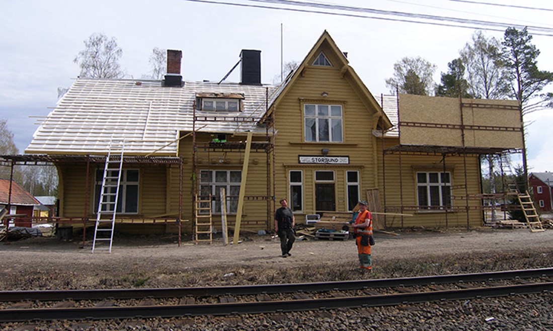 Exteriörbild av Järnvägsstationen i Storsund. Framför huset står två män, och en av dem har orange varselkläder.