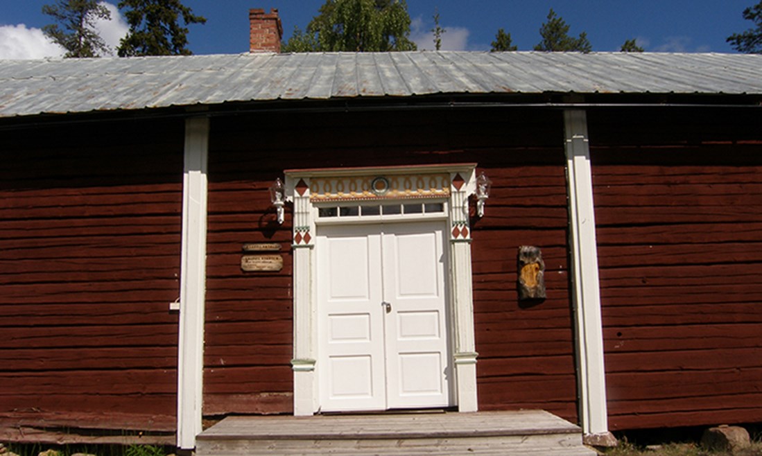 Kangos hembygdsgård, fotograferad mot väggen med entrédörren som har dubbla dörrar. Vädret är soligt.