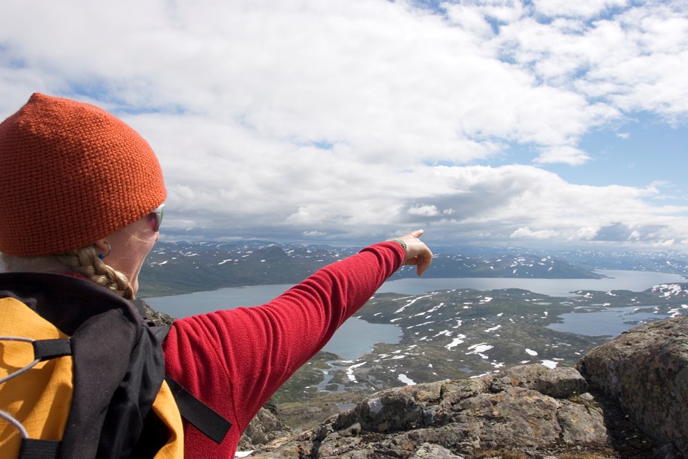 En kvinna med ryggsäck som står på ett fjäll och pekar mot sjöar och fjäll längre bort.